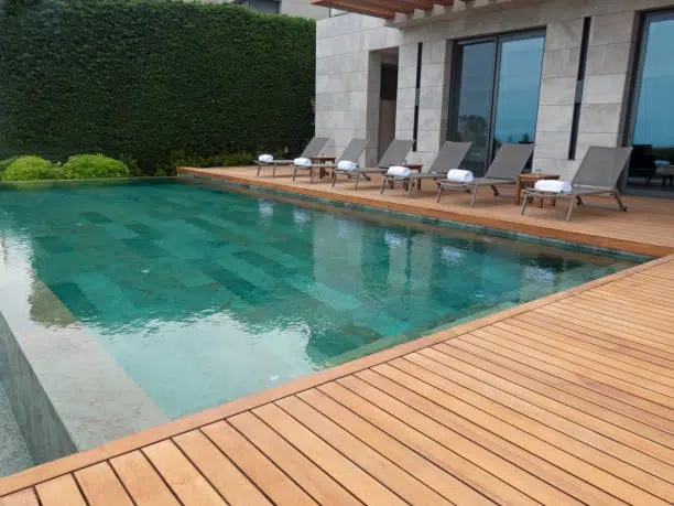 Modern pool with wooden deck, lounge chairs, and privacy hedge backdrop. -deck and pools